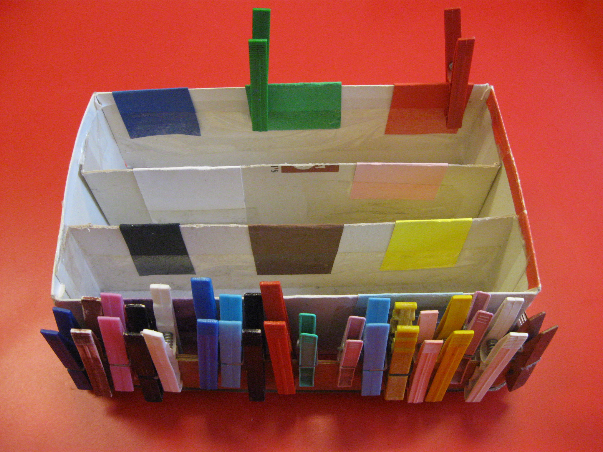 Una scatola rettangolare bianca con inserti colorati e, su un lato, mollette di diversi colori appese.