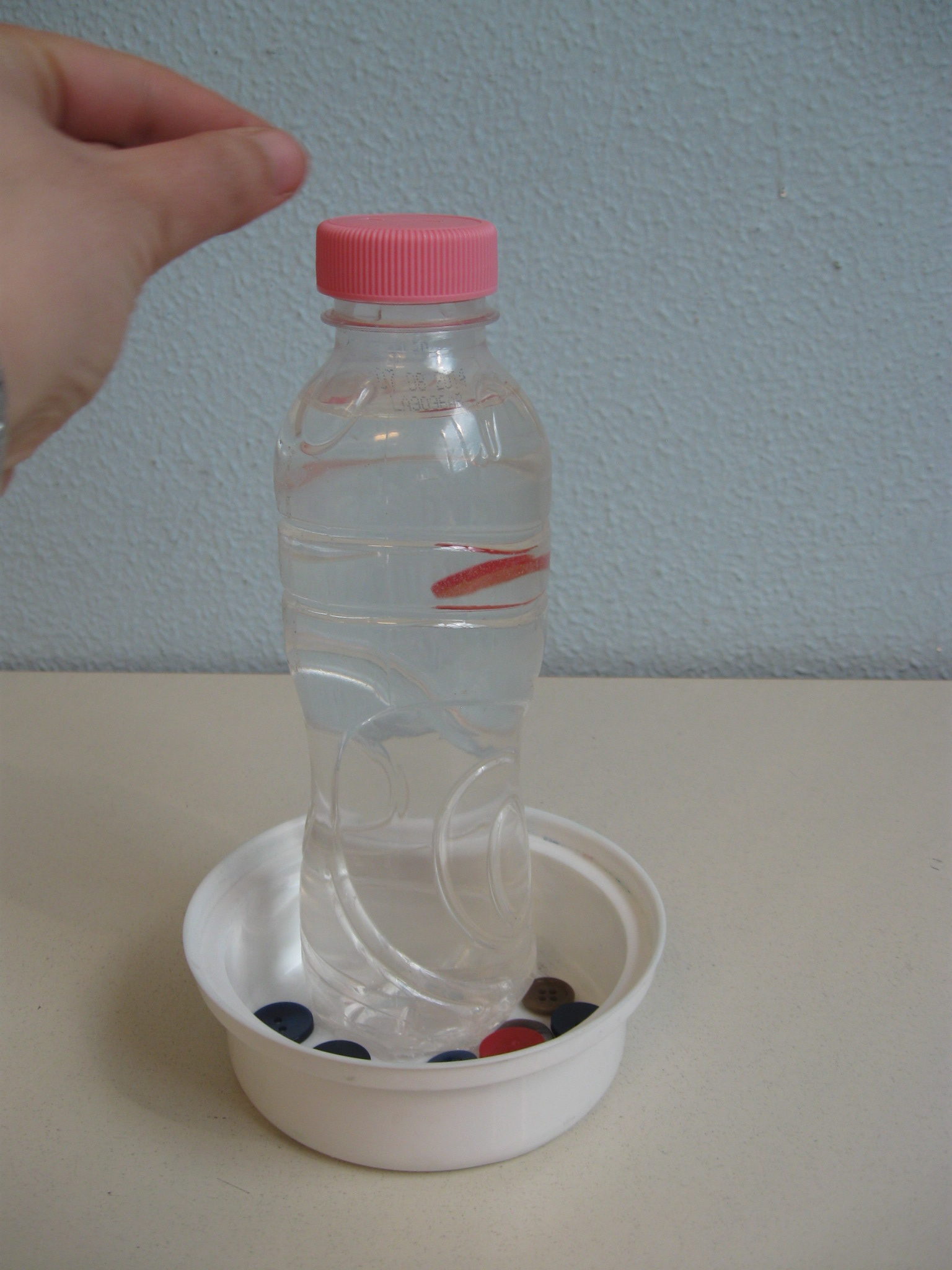 Una mano ha appena infilato un bottone nella fessura del tappo di una bottiglia: il bottono cade lentamente nell'acqua contenuta nella bottiglia.